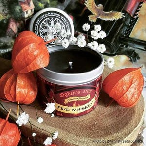 May include: A red and gold candle tin with the text "Ogden's Olde Cinnamon Flavored Fire Whiskey Est. 1860". The candle is surrounded by white flowers and orange paper lanterns.