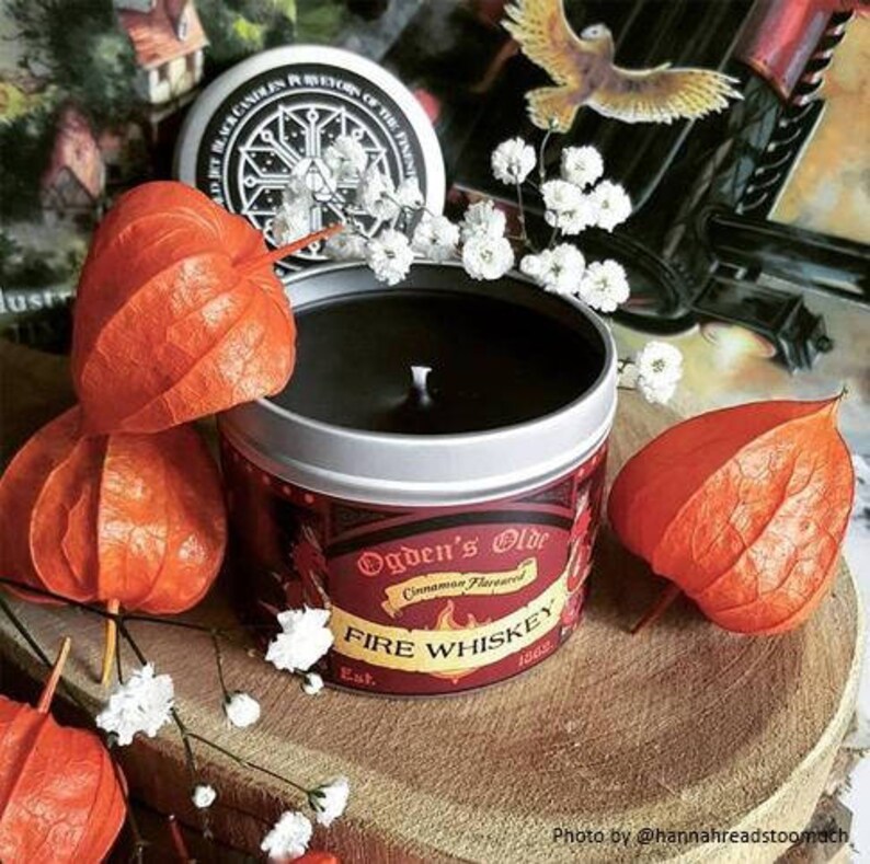 May include: A red and gold candle tin with the text "Ogden's Olde Cinnamon Flavored Fire Whiskey Est. 1860". The candle is surrounded by white flowers and orange paper lanterns.