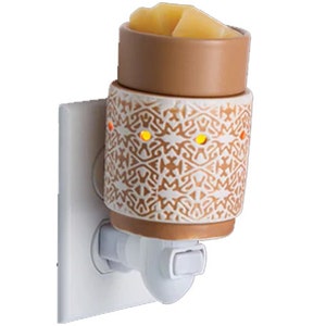 Puede incluir: Un calentador de cera enchufable blanco con un plato de cerámica de color marrón y blanco con estampado. El calentador tiene una luz que ilumina la cera derretida dentro del plato.