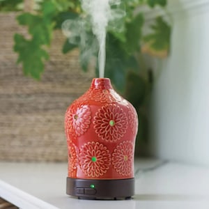 Puede incluir: Difusor de aceites esenciales de cerámica roja con un diseño floral. El difusor está sobre una superficie blanca y tiene una base marrón con una luz verde.