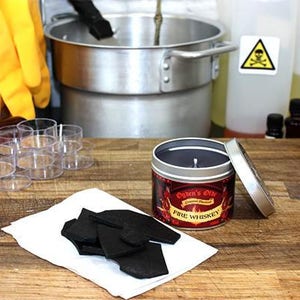 May include: A red and gold candle in a tin container with the text "Ogden's Oils" and "Fire Whiskey" on the label. The candle is surrounded by small glass containers and a white surface with black pieces of wax.