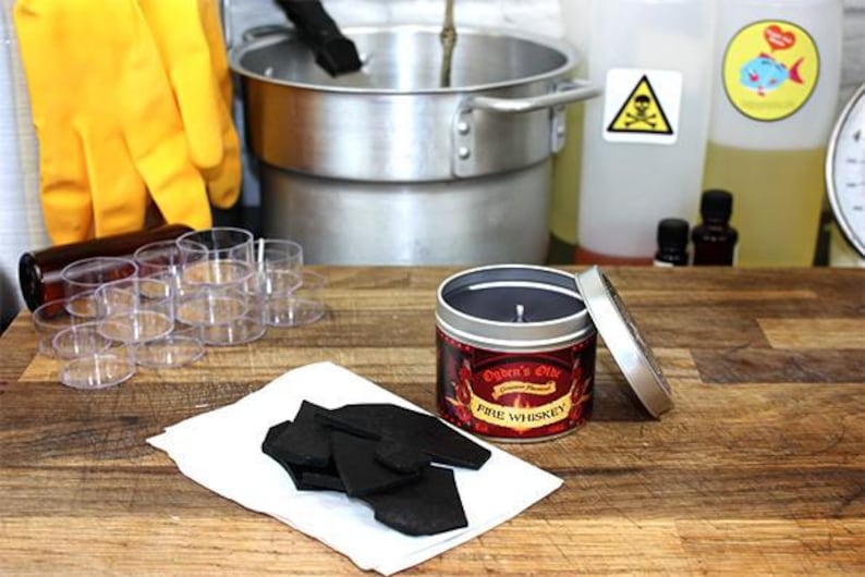 May include: A red and gold candle in a tin container with the text "Ogden's Oils" and "Fire Whiskey" on the label. The candle is surrounded by small glass containers and a white surface with black pieces of wax.