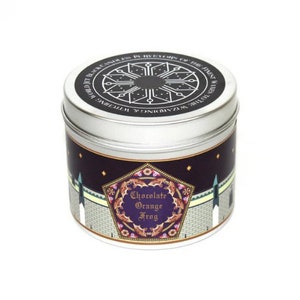 Puede incluir: Una vela en una lata de plata con un diseño en blanco y negro en la tapa. La etiqueta en la lata presenta un fondo morado con una forma de pentágono dorado. Dentro del pentágono hay imágenes de murciélagos y el texto "Chocolate Orange Frog".