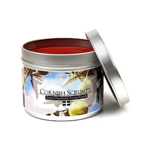 Könnte beinhalten: Eine silberne Blechkerze mit rotem Wachsoberteil und einem Etikett mit der Aufschrift "Cornish Scrumpy Artisan Handmade Scented Candle". Das Etikett zeigt ein Bild von Äpfeln und einem blauen Himmel.