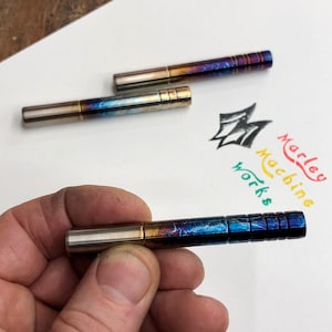 May include: Three metal pen-like objects with a blue, gold and silver finish. The objects have a textured surface and are arranged on a white surface. The text "Marley Machine Works" is visible in the background.