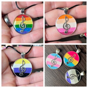 May include: Four necklaces with round pendants featuring a silver treble clef on a background of different pride flags. The flags are rainbow, pink, white, and orange, blue, pink, and white, yellow, purple, and black, and white, purple, and black.