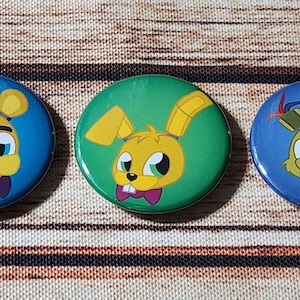 May include: Three round buttons with cartoon images of a yellow bear, a yellow bunny rabbit, and a yellow fox. The bear is wearing a purple hat and bow tie. The bunny is wearing a purple bow tie. The fox is wearing a blue and red feather. The buttons are on a wooden surface.