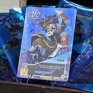 Pode incluir: Um ba&uacute; de tesouro de madeira cheio de pacotes de cartas azuis e uma &uacute;nica carta colecion&aacute;vel. A carta apresenta um personagem de desenho animado com o texto "VCH". O ba&uacute; est&aacute; em uma superf&iacute;cie arenosa.