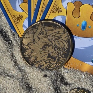 Puede incluir: Un conjunto de cartas del juego "Royalty Card Game" con un dise&ntilde;o dorado y azul, junto con una moneda de oro con una ilustraci&oacute;n detallada, sobre una superficie arenosa. Varias monedas de oro est&aacute;n dispersas alrededor de las cartas.