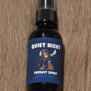May include: A dark glass spray bottle with a black cap, labeled "Quiet Night Fursuit Spray." The label features a cartoon character and the text "Fursuit Spray." The bottle contains distilled water, isopropyl alcohol, and fragrance oil.