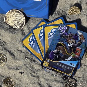 Pode incluir: Um jogo de cartas com uma caixa azul, cartas e moedas de ouro na areia. As cartas apresentam uma personagem de desenho animado com cabelo roxo e o texto "YCH". Uma moeda de ouro com um desenho de lobo tamb&eacute;m est&aacute; presente.