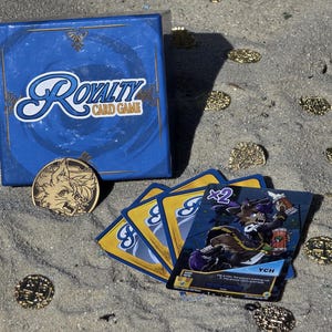 Pode incluir: Uma caixa azul e cartas do Royalty Card Game s&atilde;o exibidas na areia, juntamente com uma moeda de ouro e moedas de ouro espalhadas. A caixa tem o logotipo do jogo em escrita branca. As cartas apresentam uma ilustra&ccedil;&atilde;o de personagem.