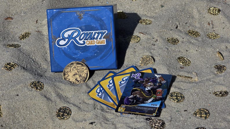Pode incluir: Uma caixa azul e cartas do Royalty Card Game s&atilde;o exibidas na areia, juntamente com uma moeda de ouro e moedas de ouro espalhadas. A caixa tem o logotipo do jogo em escrita branca. As cartas apresentam uma ilustra&ccedil;&atilde;o de personagem.