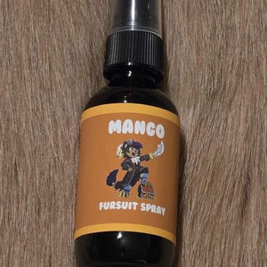 May include: A dark brown spray bottle with a black pump and a label that reads "MANGO FURSUIT SPRAY." The label features a cartoon character with a knife. The bottle is set against a brown, textured background.