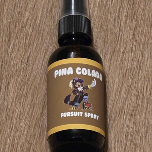 May include: A dark brown glass spray bottle with a black cap. The label reads "Pina Colada Fursuit Spray" and features a cartoon character. The bottle is on a brown, textured surface.