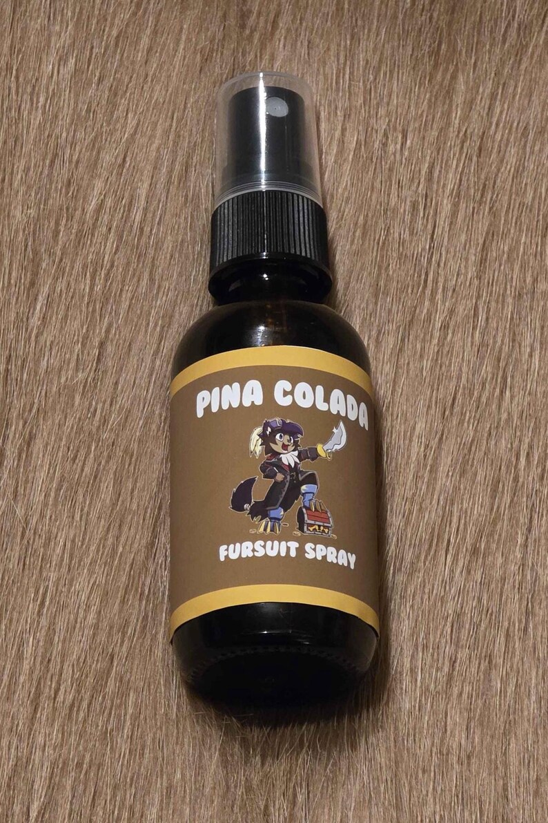 May include: A dark brown glass spray bottle with a black cap. The label reads "Pina Colada Fursuit Spray" and features a cartoon character. The bottle is on a brown, textured surface.