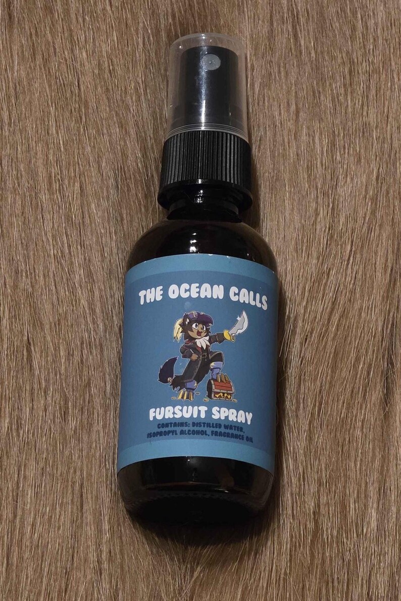 May include: A dark brown spray bottle with a black cap. The label reads "THE OCEAN CALLS FURSUIT SPRAY" with a cartoon character illustration. The bottle contains distilled water, isopropyl alcohol, and fragrance oil.