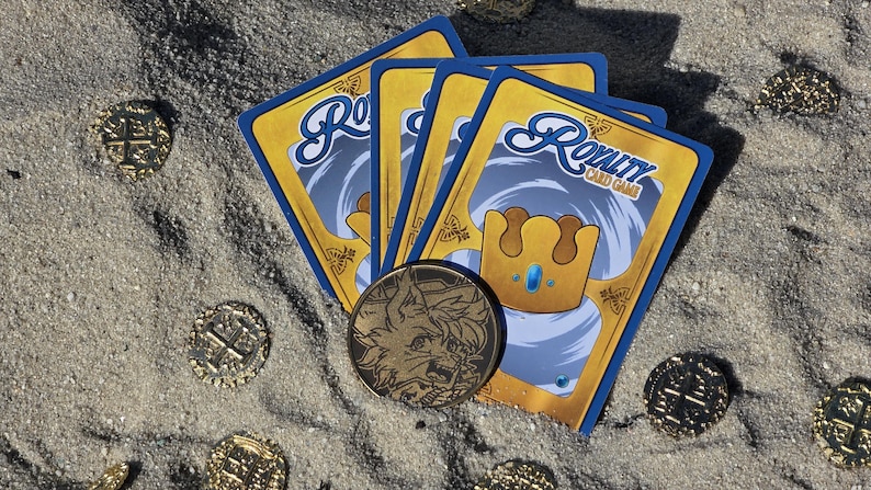 Puede incluir: Un conjunto de cartas del juego "Royalty Card Game" con un dise&ntilde;o dorado y azul, junto con una moneda de oro con una ilustraci&oacute;n detallada, sobre una superficie arenosa. Varias monedas de oro est&aacute;n dispersas alrededor de las cartas.