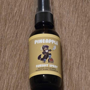 May include: A dark brown glass spray bottle with a black cap and a yellow label. The label reads "PINEAPPLE" above an illustration of a cartoon character. Below the illustration, the label reads "FURSUIT SPRAY".