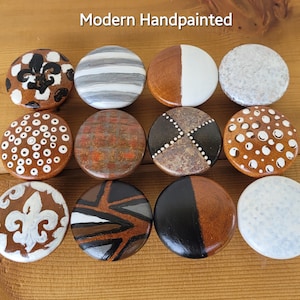 May include: Twelve hand-painted wooden knobs with various designs, including geometric patterns, dots, and a fleur-de-lis. The knobs are all brown with white and black accents. Modern Handpainted.