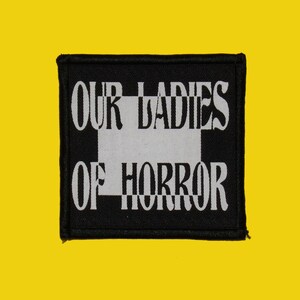 Op de afbeelding: Zwart-wit geborduurde patch met de tekst "Our Ladies of Horror" op een zwarte achtergrond.