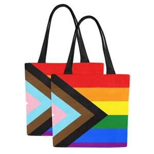 Op de afbeelding: Twee tote bags met zwarte handvatten. De tassen zijn gemaakt van canvas en hebben een regenboog pride-vlag met een zwarte en bruine driehoek in de hoek.