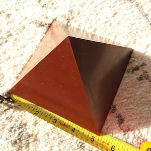 May include: A brown wooden pyramid with a measuring tape along one side. The pyramid is approximately 15 cm in length.