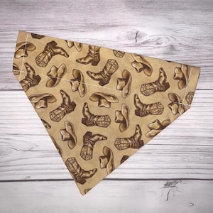 Puede incluir: Un pañuelo de color beige con un patrón de botas de vaquero y sombreros marrones. El pañuelo tiene forma triangular y un borde cosido blanco.