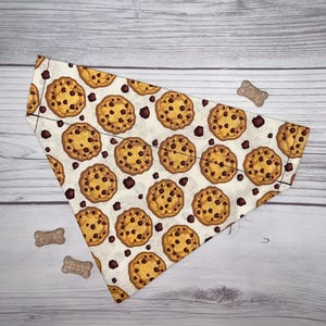 以下が含まれることがあります： オフホワイトの背景に黄金色のチョコチップクッキーの繰り返し模様がプリントされた三角形のペット用バンダナ。布製アクセサリーの周囲には、骨の形をした小さな犬用おやつが3つ配置されています。