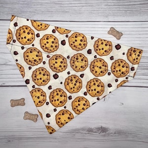 May include: A white dog bandana with a pattern of chocolate chip cookies. The bandana is triangular in shape and has a white background with brown chocolate chip cookies.