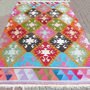May include: A rectangular area rug featuring a repeating diamond pattern in vibrant colours including red, pink, orange, green, blue, and dark brown. The design incorporates white geometric motifs within each diamond, finished with white fringe.