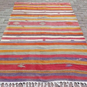 May include: A rectangular woven rug featuring horizontal stripes in shades of red, orange, yellow, purple, and white. The rug includes small, scattered geometric embroidered motifs throughout the surface and is finished with white fringe at both ends.