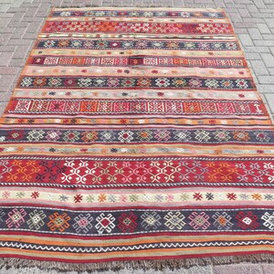 May include: A rectangular woven rug featuring horizontal bands of geometric patterns in shades of red, orange, dark blue, and cream. The design includes repeating diamond and cross motifs across the surface, finished with a short fringe at the ends.