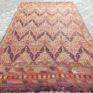 May include: A rectangular woven rug featuring a repeating zigzag pattern in shades of red, orange, yellow, and dark brown. The design includes intricate geometric motifs within the zigzags and a wide horizontal border with fringe at the bottom edge.