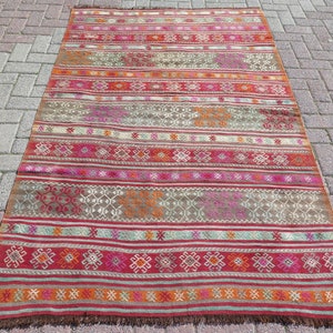 May include: A rectangular woven rug featuring horizontal bands of red, orange, pink, and brown. The design includes repeating geometric patterns and thin stripes throughout the surface, finished with a short brown fringe along the bottom edge.