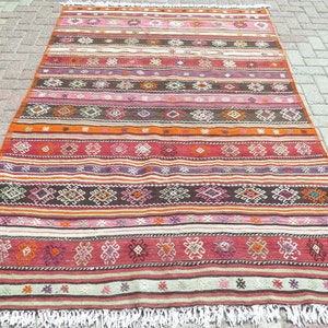May include: A rectangular woven rug featuring horizontal stripes in shades of red, brown, orange, pink, and white. The rug is decorated with repeating geometric diamond and floral motifs across the surface and finished with white fringe at both ends.