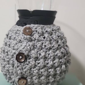 May include: A gray, crocheted coffee maker cozy with three brown buttons. The cozy is wrapped around a glass coffee carafe with a black collar. The cozy is designed to keep coffee warm.