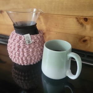 Può includere: Un copri-caffè in crochet rosa con un'etichetta che recita "Owl Hook Up Creations" è avvolto attorno a una caraffa per caffè in vetro. Una tazza in ceramica verde chiaro con un manico nero è piena di caffè e si trova su una superficie nera.