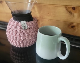 May include: A pink crocheted coffee cozy with a tag that reads "Owl Hook Up Creations" is wrapped around a glass coffee carafe. A light green ceramic mug with a black handle is filled with coffee and sits on a black surface.