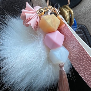 以下が含まれることがあります： ピンクの革ひも、金色のリング、ピンクと白のビーズチャームが付いた、白いふわふわのポンポンキーホルダー。チャームにはピンクのタッセルと金色のビーズが付いています。