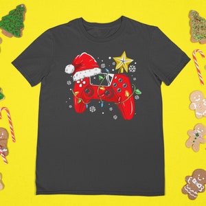 May include: Dark gray t-shirt featuring a red video game controller decorated with a Santa hat, Christmas lights, and a gold star. The shirt is surrounded by gingerbread cookies, candy canes, and Christmas trees on a yellow background.