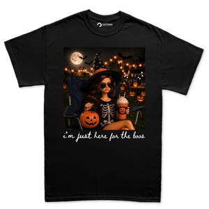 May include: A black t-shirt featuring a graphic of a figure wearing a witch hat and sunglasses, holding a pumpkin bucket and a drink. The figure wears a skeleton-print top. Below the graphic, the white text reads: i'm just here for the boos.