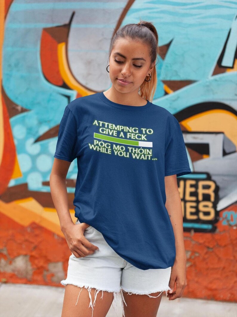 A young woman wearing a blue t-shirt with a humorous text printed on it, standing in front of a colorful graffiti wall.