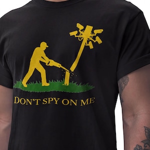 May include: A black t-shirt featuring a yellow graphic of a figure using a chainsaw to cut down a utility pole topped with multiple surveillance cameras. The graphic includes a patch of green grass and the text DON'T SPY ON ME in yellow letters.