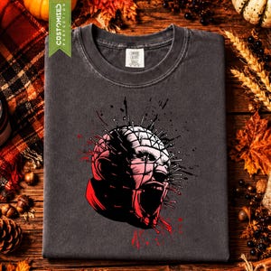 May include: A charcoal gray crew neck t-shirt featuring a graphic illustration of a head covered in nails, with red and black ink splatter accents. The design depicts a stylized character with a grid pattern on the skin and an open mouth.