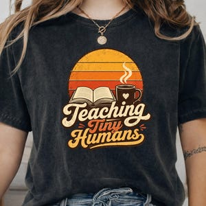 Può includere: Una t-shirt grigio antracite con la grafica di un libro aperto e una tazza fumante con un cuore, su uno sfondo di tramonto retro arancione e giallo. Il testo "Teaching Tiny Humans" è stampato in un carattere corsivo color crema.