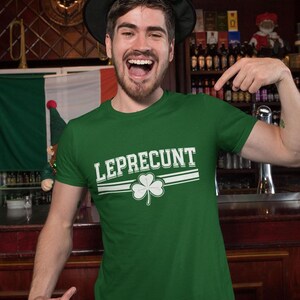 A man wearing a green t-shirt with the word "LEPRECIUNT" and a green hat is smiling and pointing at the camera in a bar setting.
