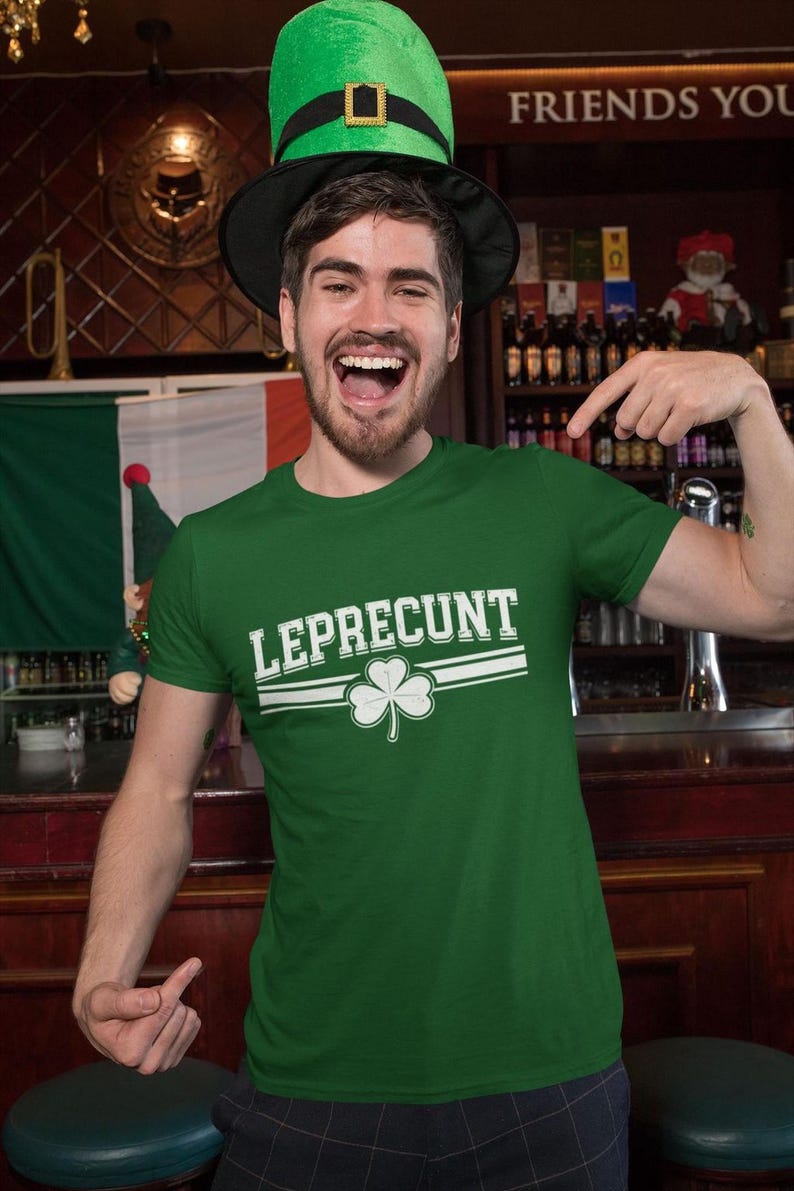 A man wearing a green t-shirt with the word "LEPRECIUNT" and a green hat is smiling and pointing at the camera in a bar setting.