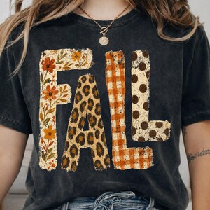 Puede incluir: Una camiseta gris carbón con la palabra FALL en letras grandes. Cada letra tiene un patrón distinto: la F tiene flores naranjas y amarillas, la A tiene un estampado de leopardo marrón, la L tiene un cuadro naranja y la L tiene puntos marrones.