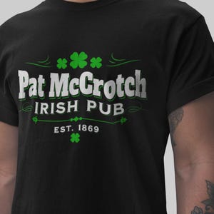 Op de afbeelding: Zwart T-shirt met de tekst "Pat McCrotch Irish Pub" in wit en groen. Het ontwerp bevat klavers en het oprichtingsjaar 1869. Een casual T-shirt voor St. Patrick's Day of dagelijks gebruik.